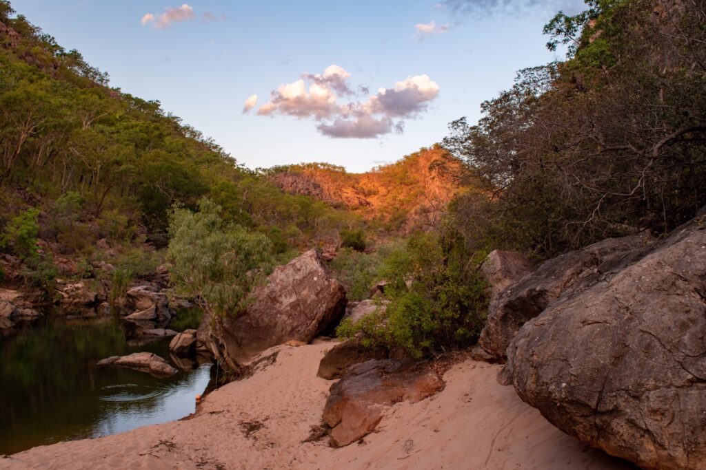 A small waterhole with a sandy beach and the sunset turning a rocky escarpment red in the distance