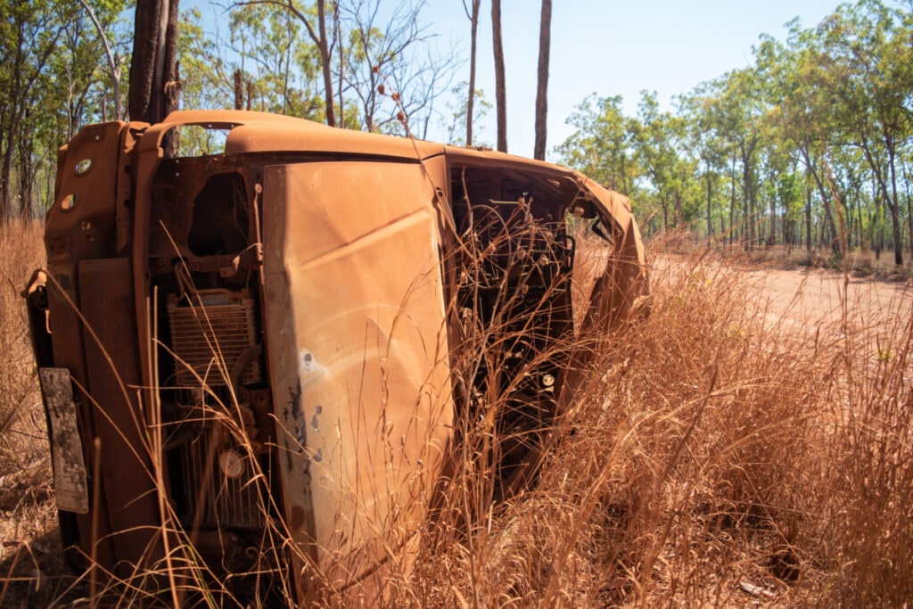 A old wrecked car lies on its side covered in red dust alongside a dirt road