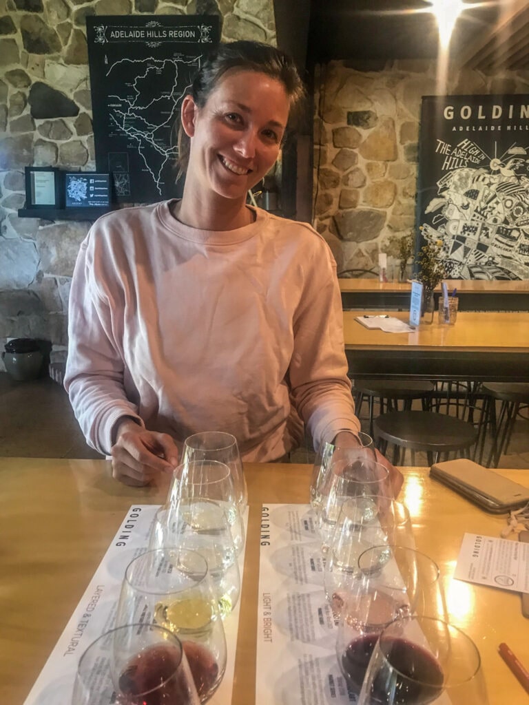A woman in a pink sweater smiles for the camera sitting in front of a couple of wine flights at a winery