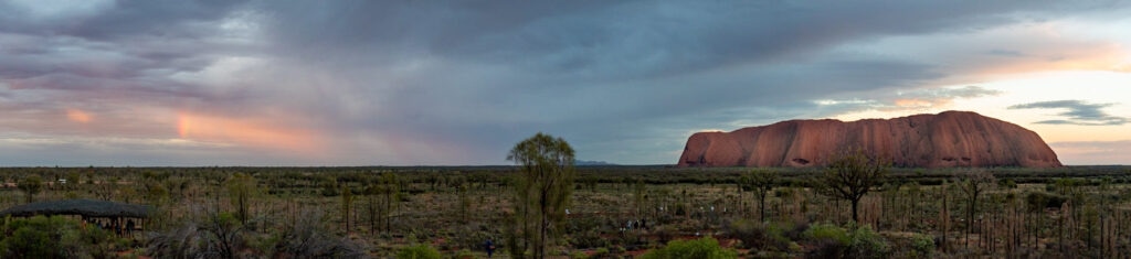 A panorama of Uluṟu/Ayers Rock with rain clouds and grassy foreground