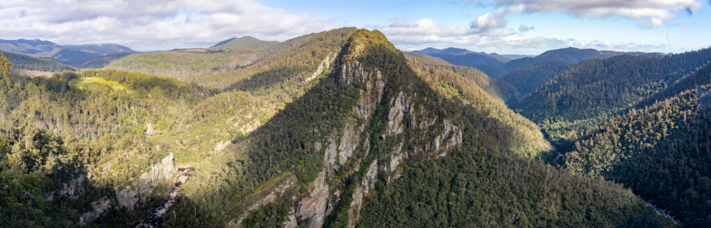 A panoramic photo of Leven Canyon, Tasmania, Australia, with rocky outcrops covered in green trees and more mountains in the horizon on a partly cloudy day