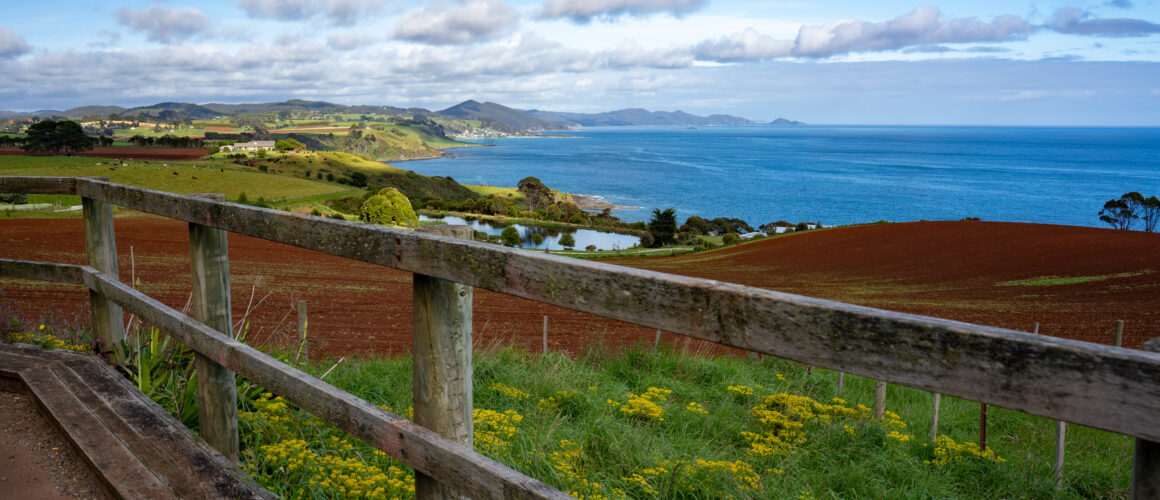 A picture of a fence and yellow wildflowers in the foreground, rolling red and green farmland and mountain peaks in the distance, and blue ocean stretching into the horizon on a partly cloudy day in Northwest Tasmania, Australia