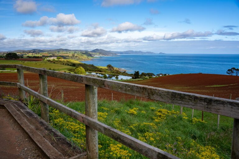 A picture of a fence and yellow wildflowers in the foreground, rolling red and green farmland and mountain peaks in the distance, and blue ocean stretching into the horizon on a partly cloudy day in Northwest Tasmania, Australia
