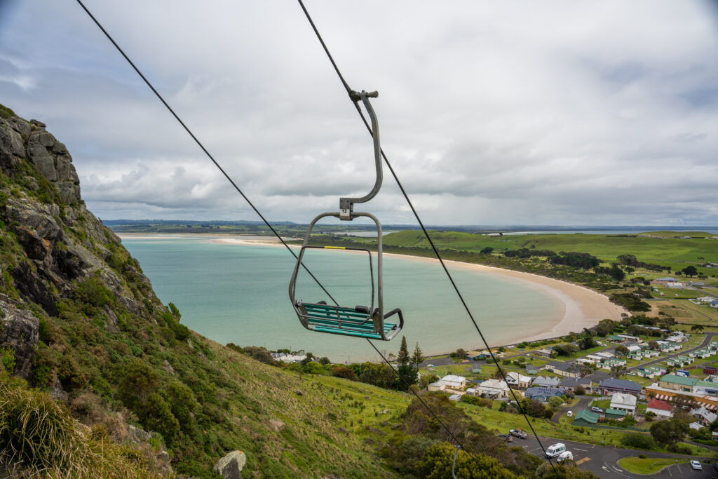 An empty chair on the chairlift heading down from the Nut in Stanley, Tasmania, Australia, of heathland and in the distance a small town, a blue ocean and a white sand beach stretching away from rolling farmland on a cloudy day