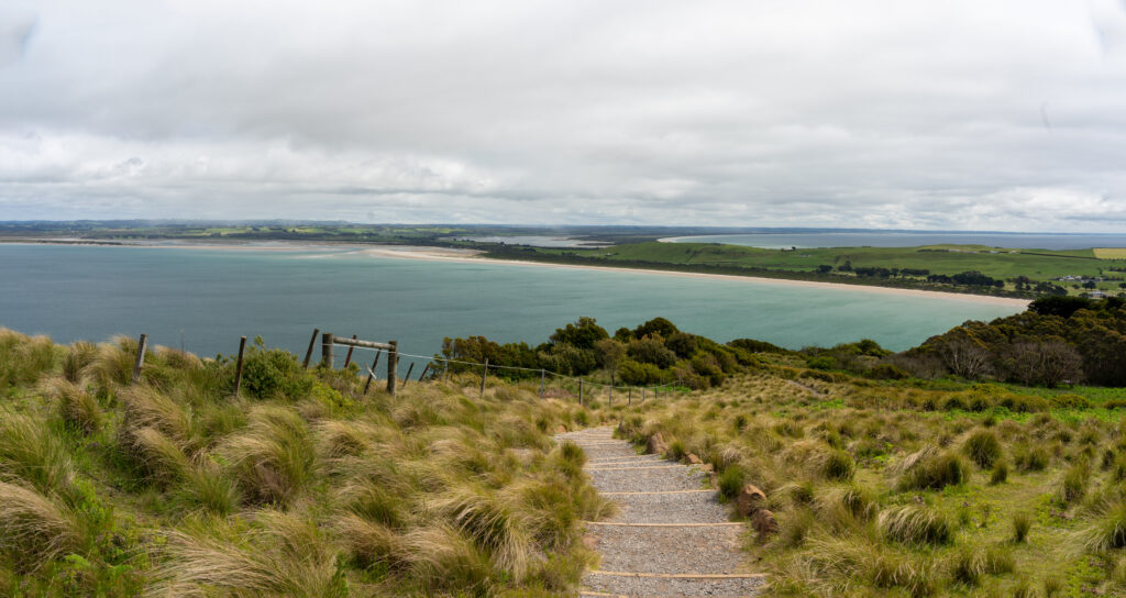 A panoramic view from the top of The Nut in Stanley, Tasmania, Australia, of heathland and in the distance ocean and a white sand beach stretching away from rolling farmland on a cloudy day