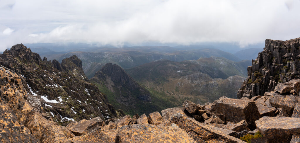 A view from high up on the summit of Cradle Mountain of rocky outcrops and more peaks in the distance and low lying cloud in Cradle Mountain National Park, Tasmania, Australia
