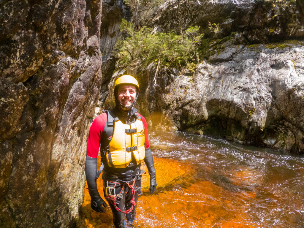 A man in canyoning gear smiles at camera as he stands next to waterway in Dove Canyon, Cradle Mountain, Tasmania, Australia