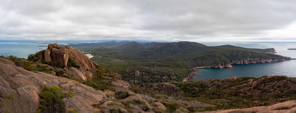 A panoramic view from atop granite rocks rolling down green hills to a larger landmass surrounded by ocean on a cloudy day