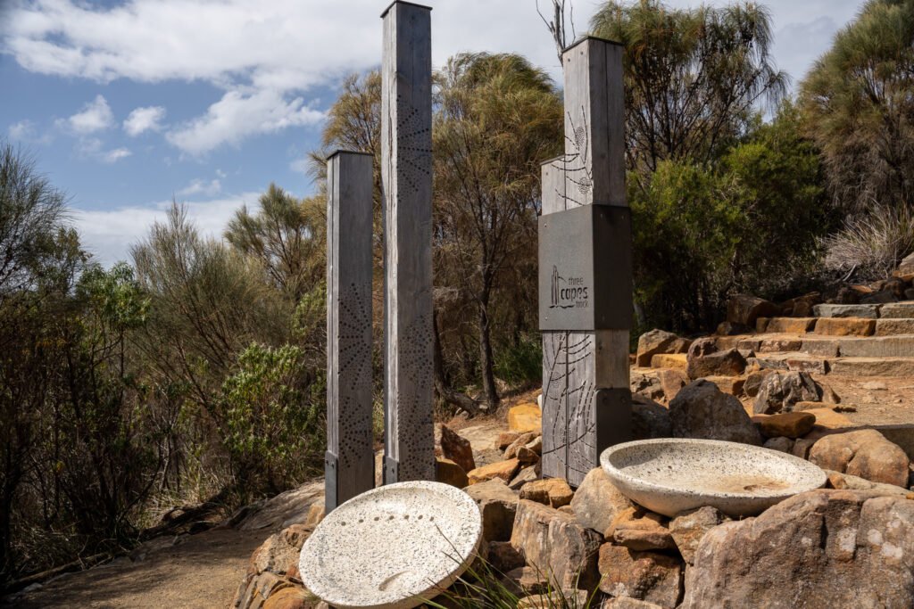 A sign for the Three Capes Track, with 3 wooden pillars and 2 stone shallow bowls, with steps leading out of frame on a partly cloudy day