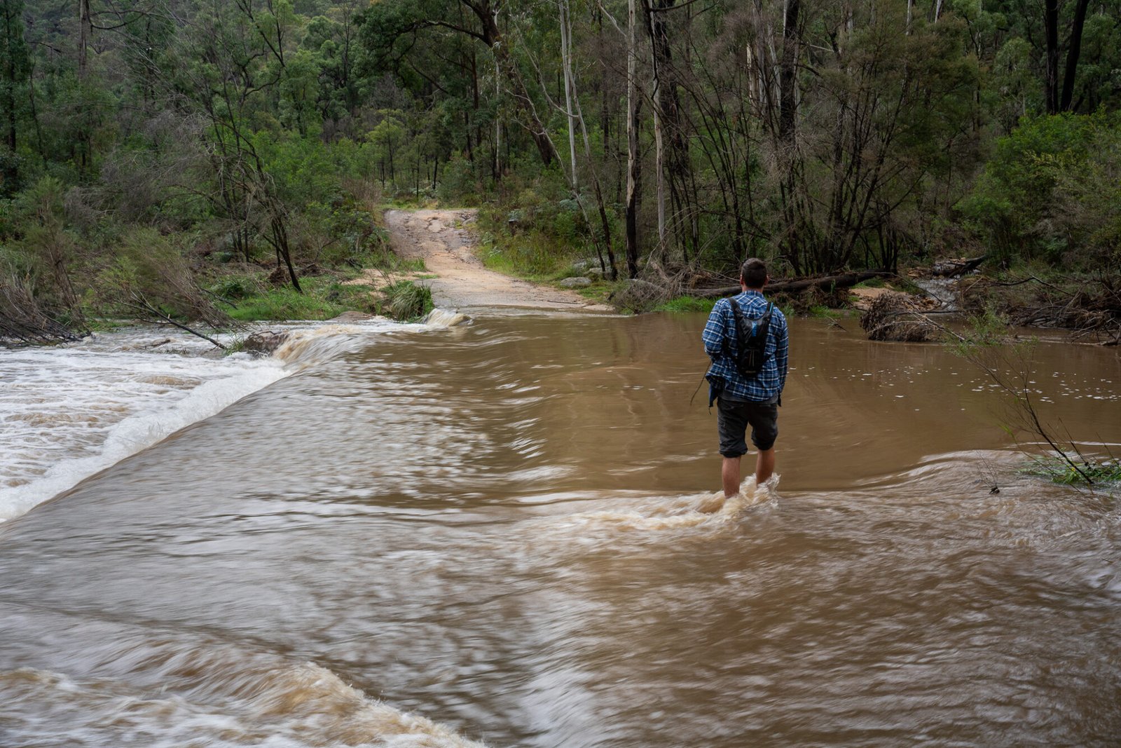 A man wearing a blue flannelette shirt and long dark pants with a Camelbak has his pant legs roll up as he walks through fast moving water to reach a dirt track on the other side