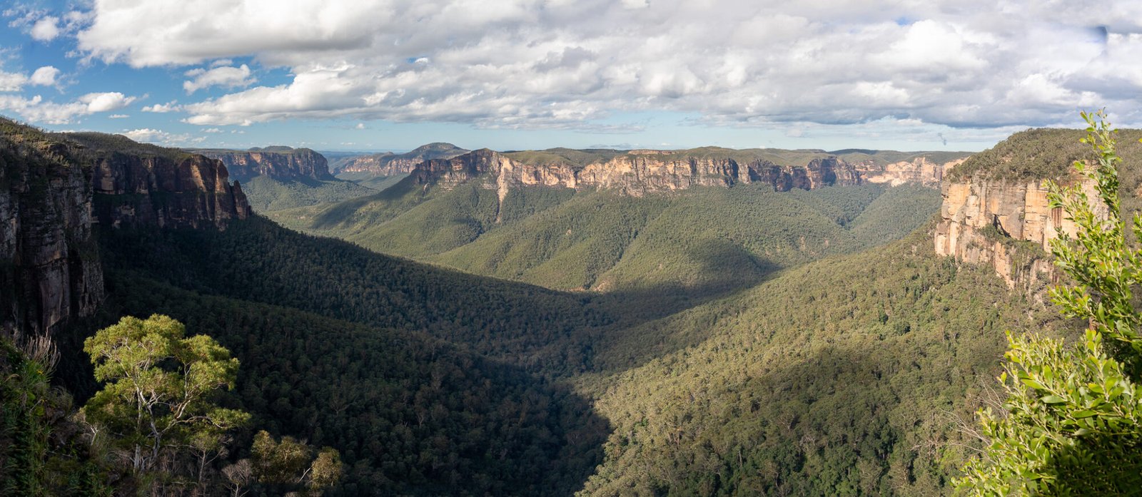 A panoramic view of rocky cliffs dropping dramatically into a treed valley half covered in shade on a partly cloudy day