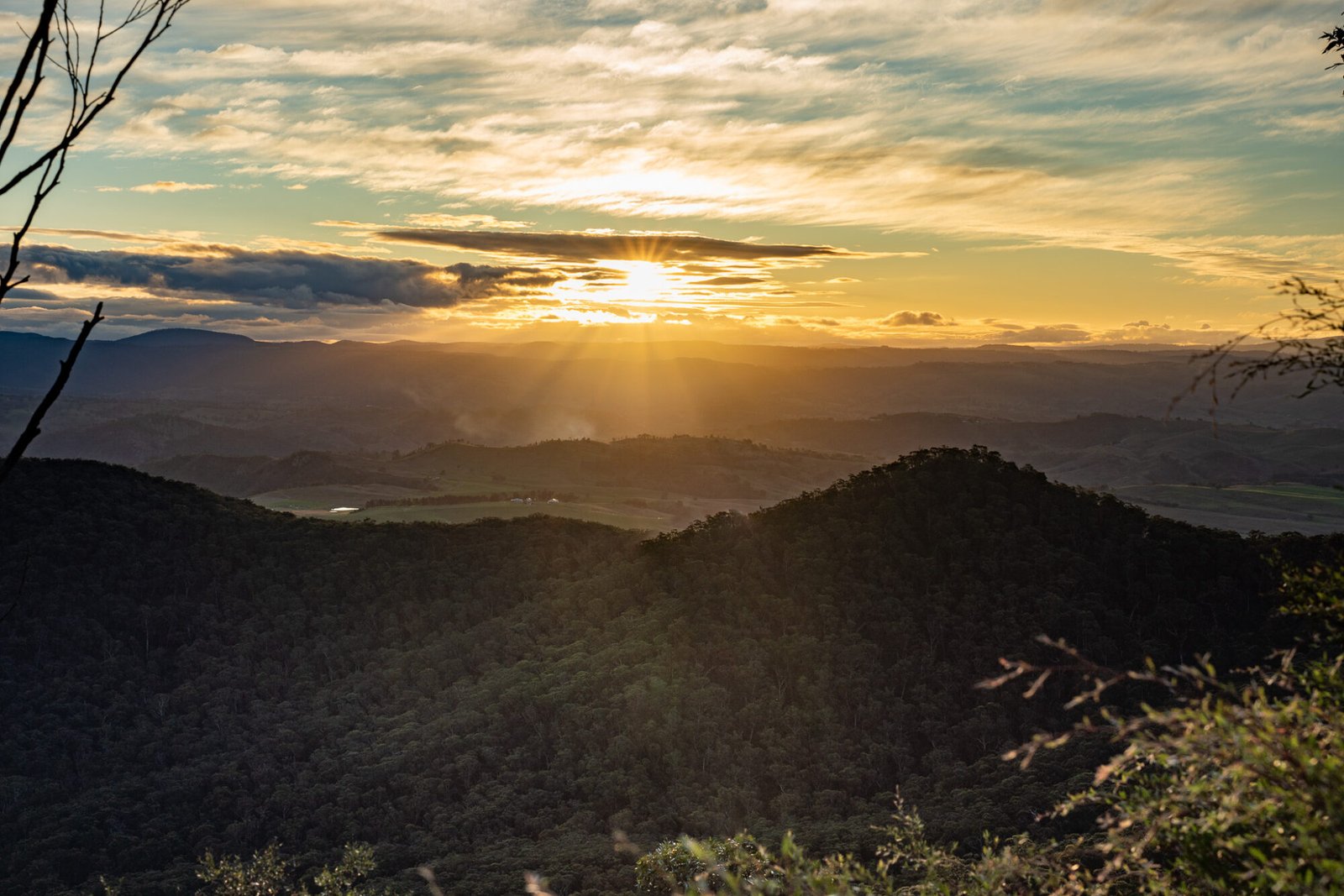 A view of rolling treed hills on a partly cloudy evening with sunbeams peaking through the clouds at sunset