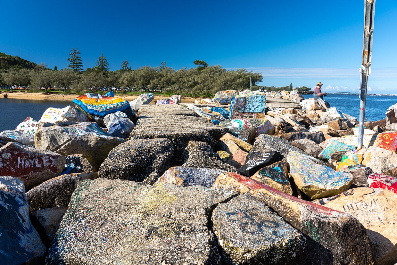 A sea rock wall with graffiti on it stretches into the background of beach and trees on a clear sunny day
