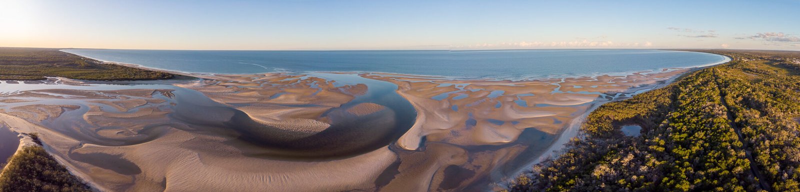 A panoramic drone shot of sandy tidal flats flanked by thick bushland looking out over the ocean at sunset