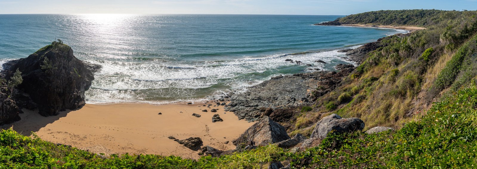 A Panoramic view of a small beach flanked by rock with a headland curving around in the background
