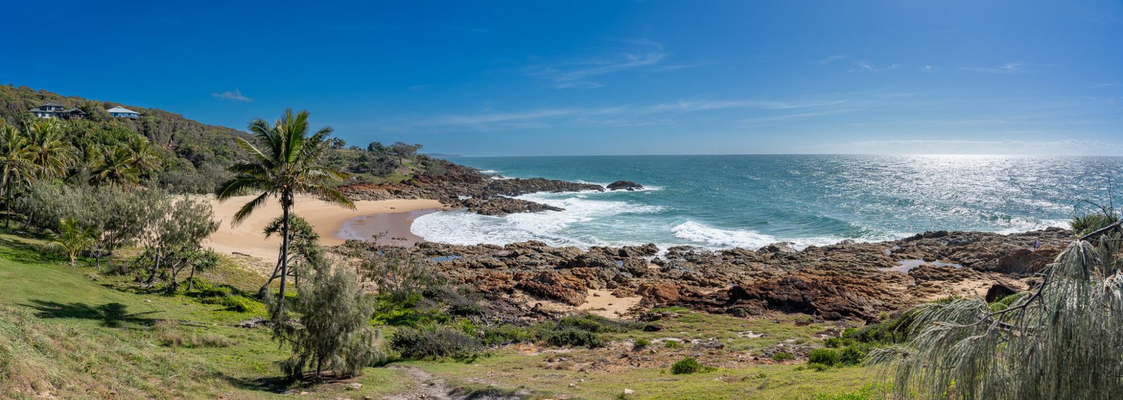 a panoramic view of grassy hils with palm trees leading to red rocks and a sandy beach overlooking a blue ocean on a clear day