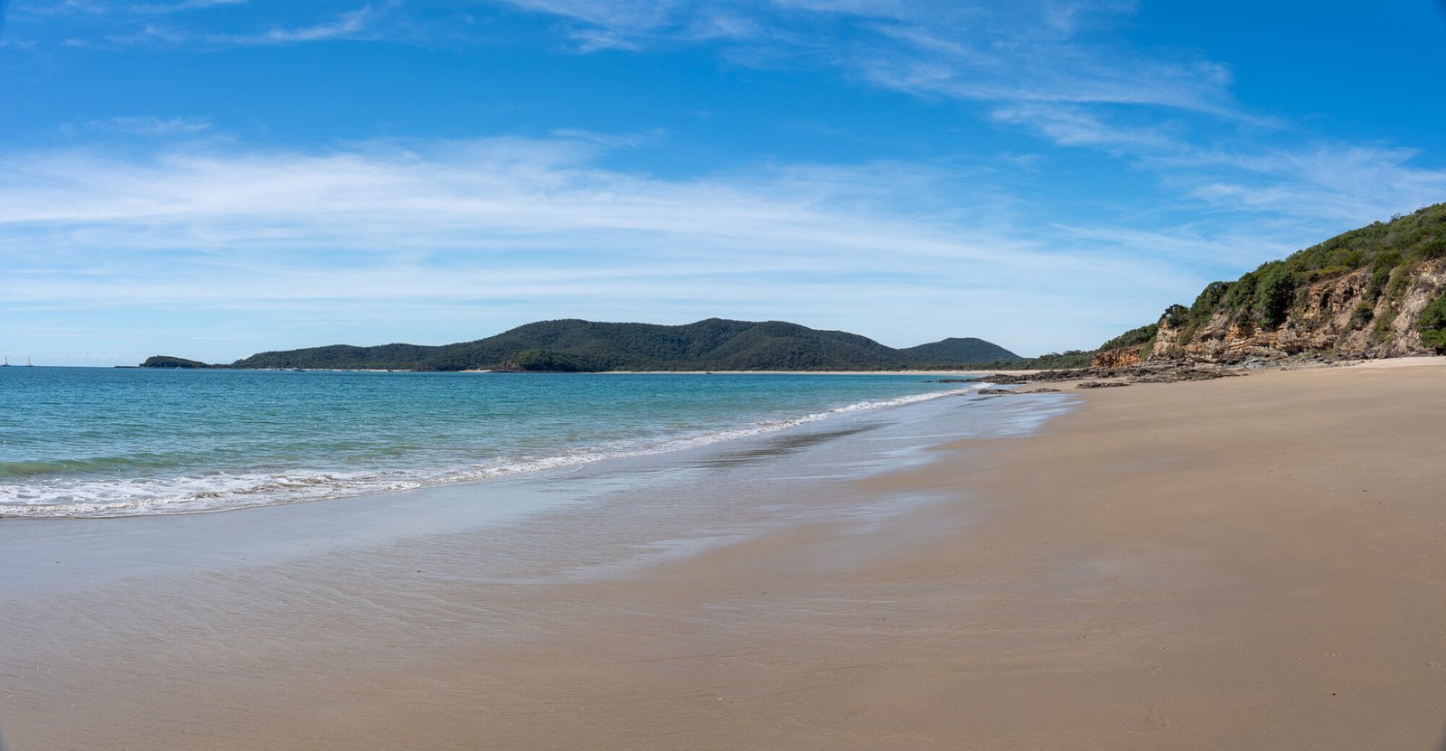 A clear sandy beaches stretches out into the distance and green rolling headlands on a partly cloudy day