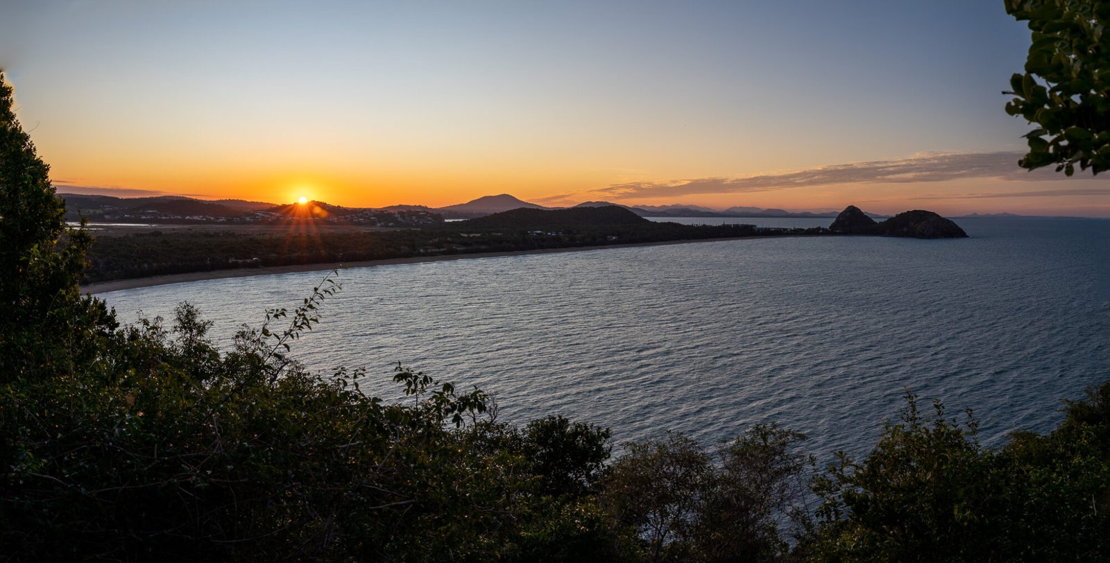 A panoramic view from high looking across the ocean to a sun setting behind tall hils