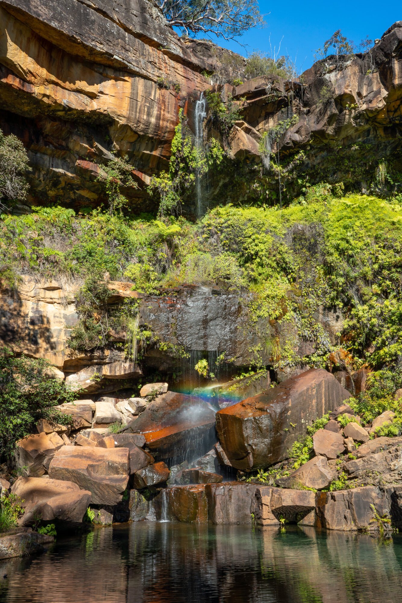 A tall rocky waterfall with a slight trickle creates a small rainbow as it lands into a reflective pool