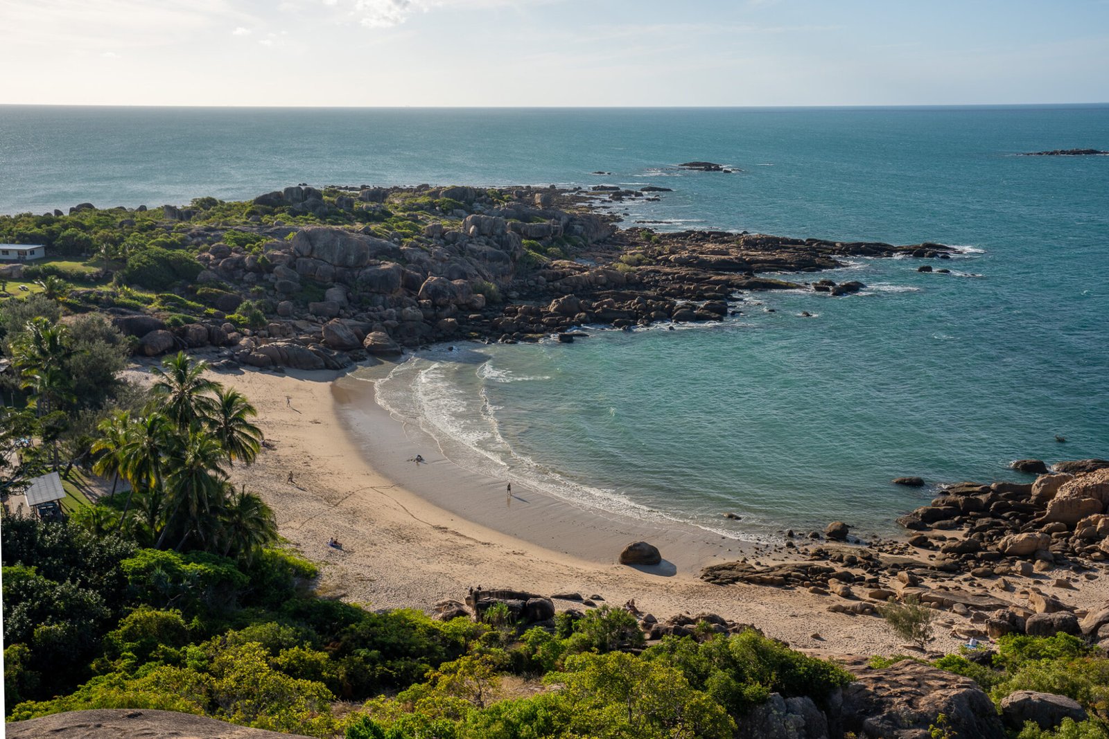 A view from high of lush vegetation leading to a small semi circular beach flanked by granite rocks looking out onto the ocean on a clear sunny day