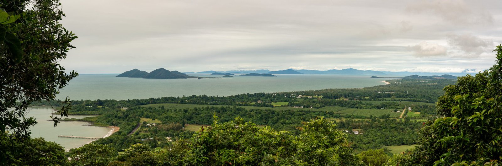 A panoramic view of above a small coastal town with lush green vegetation looking out onto numerous islands on an overcast day