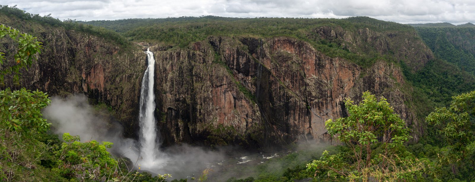 A panoramic view of a cliff face with a large single drop waterfall into a gorge with dense green vegetation all around on an overcast day