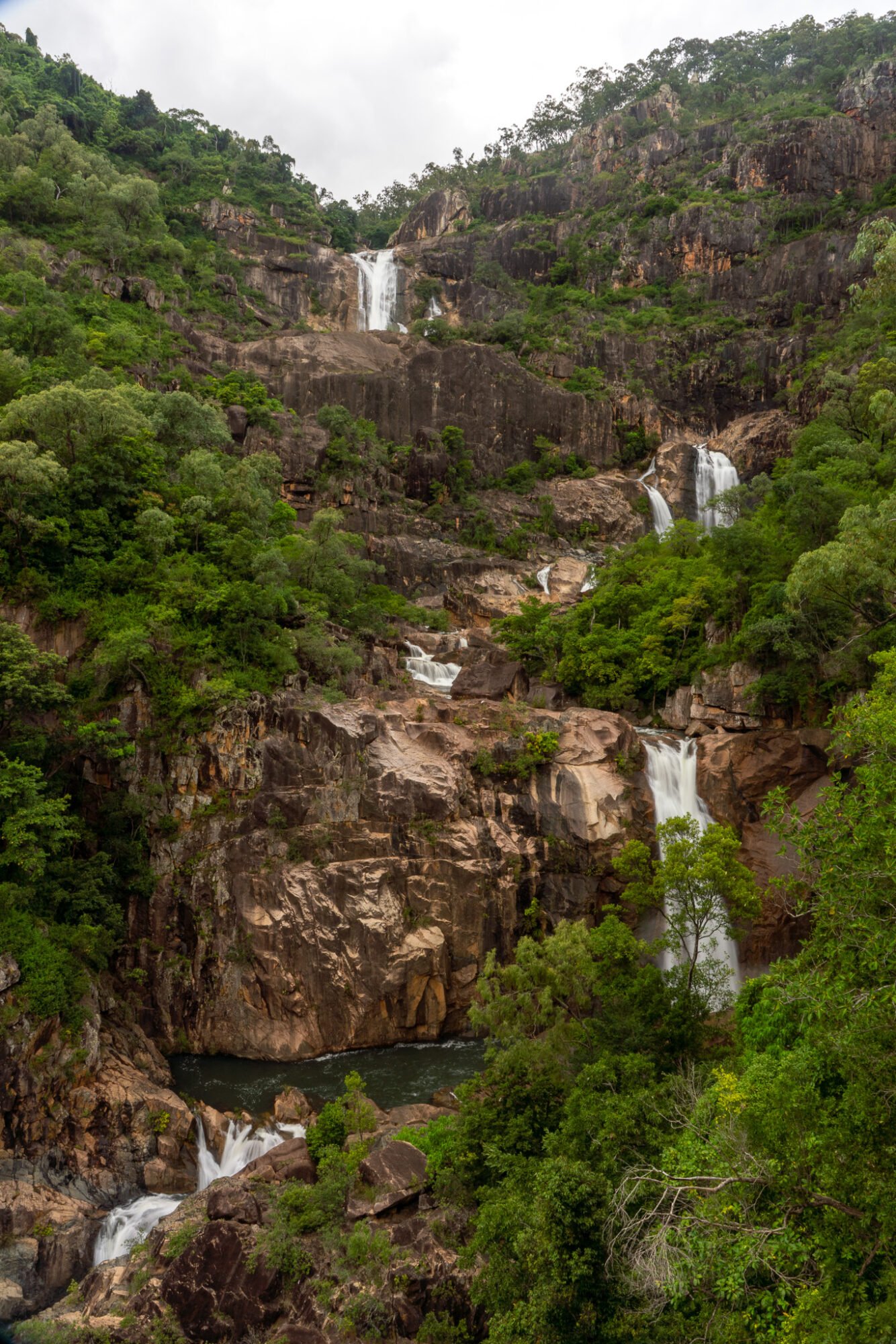 A multi tiered waterfall of granite rock framed by green vegetation on an overcast day