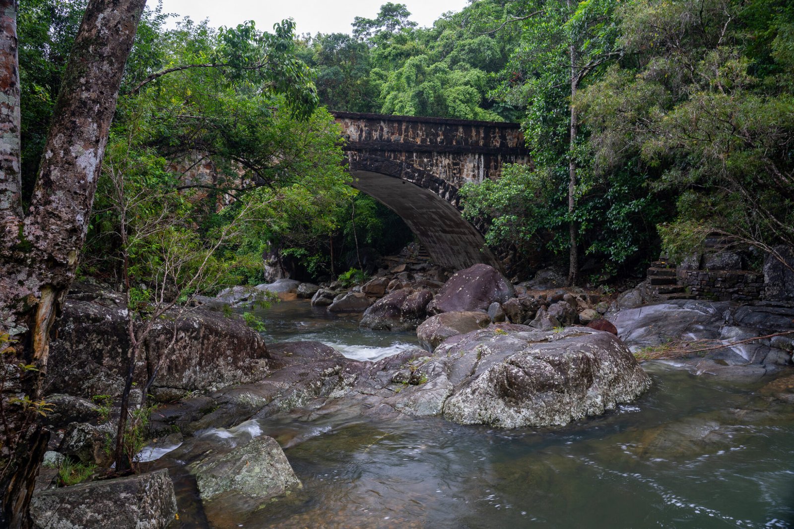 An old stony bridge surrounded on either end by lush green vegetation stands over a small creek with large boulders throughout on an overcast day