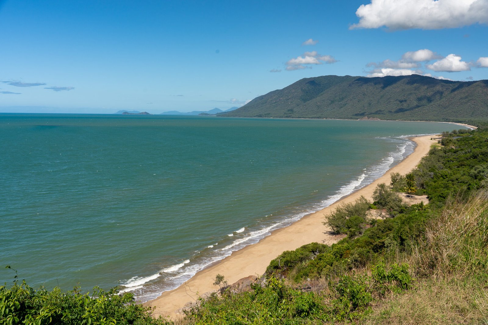 A lookout overlooking thick vegetation down to a clear sandy beach into the ocean with tall headlands in the distance on a mostly clear day