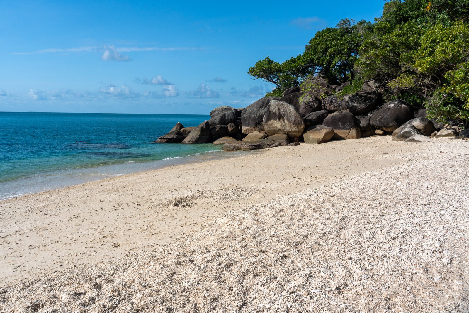 A white sandy and shelly beach leads to the ocean with granite boulders and lush green vegetation on a mostly clear day