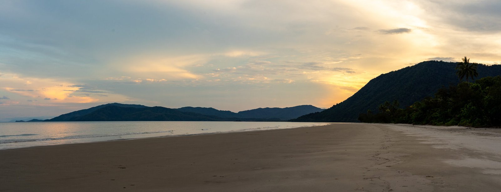 A sunset behind a large headland over clear sandy beach on a mostly cloudy evening