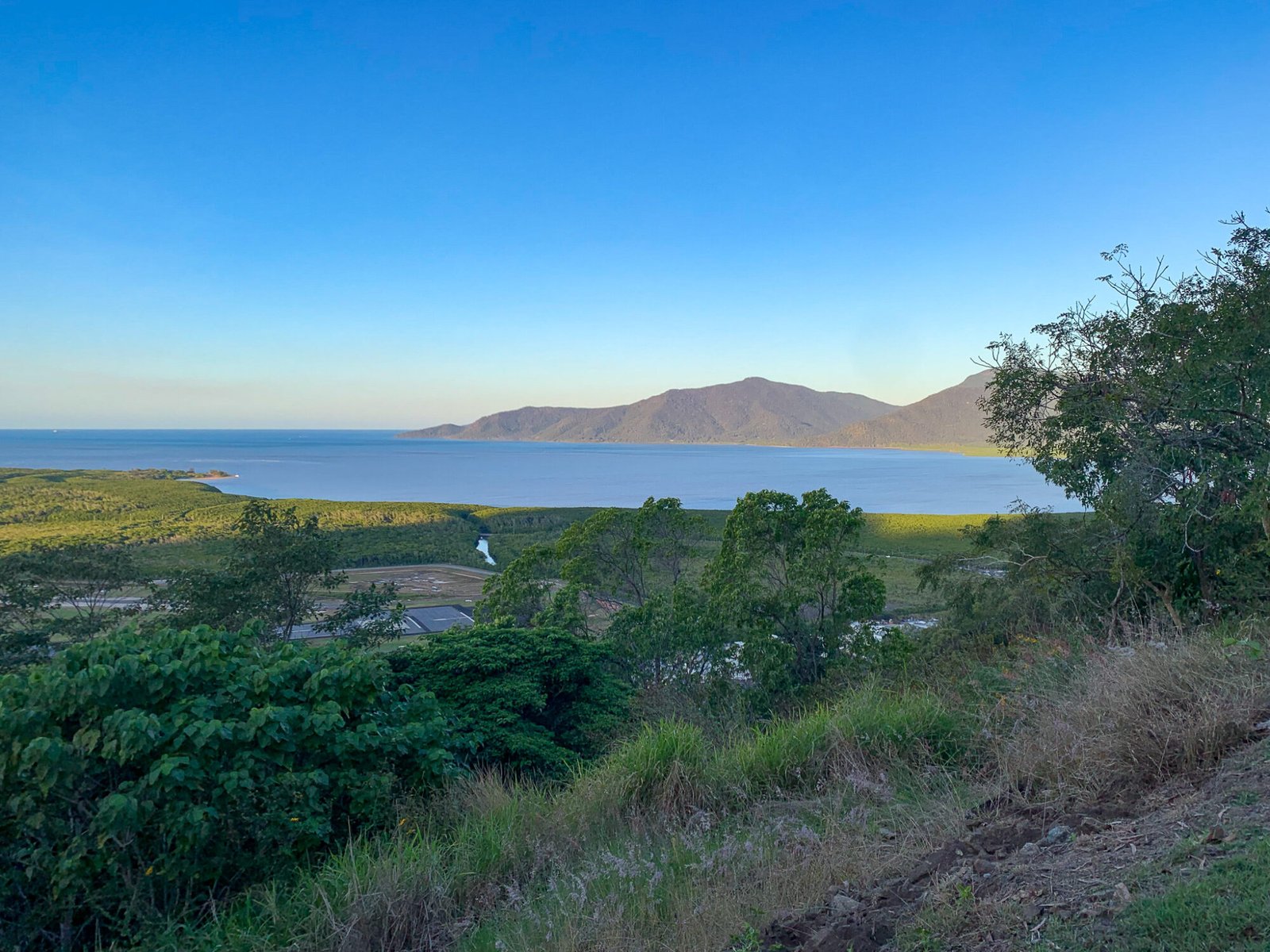 A lookout from the top of a grassy hill overlooks some town infrastructure out to the ocean and tall headlands in the distance on a clear sunny day